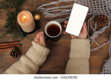 girl holds a black mug with tea, a candle burns on a wooden table, smart phone with blank screen, knitted shawl, coniferous branches and cinnamon, concept winter mood, winter is love, copy space