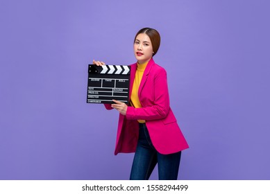 Junges schönes asiatisches Frauenmodell in der bunten Kleidung, die Filmklappe lokalisiert auf purpurrotem Hintergrund hält
