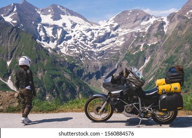 Wanita dalam pakaian penunggang basikal penuh, kura-kura pelindung jaket. Motosikal pelancongan dengan beg besar. Puncak bersalji di pergunungan Alpine di latar belakang. Laluan Grossglockner Hochalpenstrasse. Austria, Itali