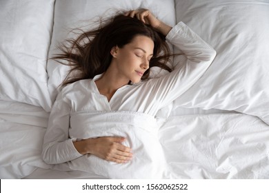 Friedliche, ruhige, schöne junge Dame trägt Pyjamas, die im gemütlichen weißen Bett auf einem weichen Kissen schlafen, das mit einer Decke bedeckt ist und ein gutes, gesundes Schlafkonzept genießt, über der Draufsicht
