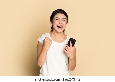 Excited happy Indian girl celebrating online win, holding phone, looking at camera isolated on brown background, overjoyed successful lucky young woman rejoicing victory, triumph, laughing