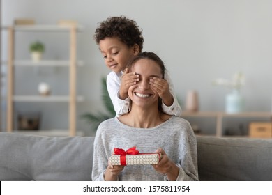 Người phụ nữ vui vẻ phấn khích ngồi trên chiếc ghế dài trong phòng khách với đôi mắt cậu bé mầm non tinh nghịch, tay cầm hộp quà được gói lại, cảm thấy tò mò. Khái niệm lễ kỷ niệm sinh nhật hoặc ngày của mẹ.