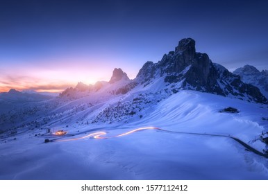 Montañas nevadas y faros de camión borrosos en el sinuoso camino por la noche en invierno. Hermoso paisaje con rocas cubiertas de nieve, casa, carretera de montaña, cielo estrellado azul al atardecer en Dolomitas, Italia