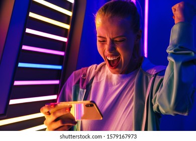 Image of happy young caucasian woman playing video games on smartphone by multicolored neon lights indoors