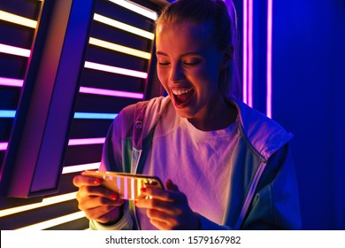 Image of beautiful young caucasian woman playing video games on smartphone by multicolored neon lights indoors