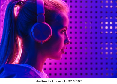 Image closeup of beautiful young caucasian woman listening to music with headphones over purple neon illumination indoors