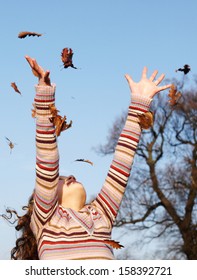 Young girl throwing dry autumn leaves up into the blue sky with her arms and and with floating leaves falling against a bright blue sky and leafless fall trees, outdoors.