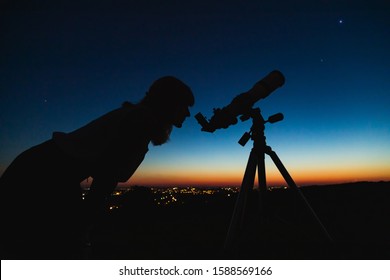 Ahli astronomi dengan teleskop melihat bintang dan Bulan dengan lampu bandar kabur di latar belakang.