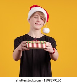 Un joven con un sombrero rojo de Papá Noel sostiene una caja de regalo y dinero en sus manos y posa en un fondo amarillo