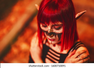 Girl cosplay in the image of a dark elf in a red wig portrait close-up. A woman with red lenses in her eyes and red hair in an elf costume with false ears and makeup. Dark elf outfit for Halloween.