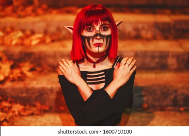 Girl cosplay in the image of a dark elf in a red wig portrait close-up. A woman with red lenses in her eyes and red hair in an elf costume with false ears and makeup. Dark elf outfit for Halloween.
