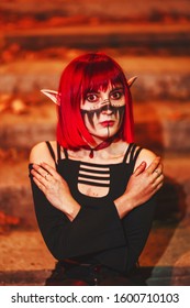 Girl cosplay in the image of a dark elf in a red wig portrait close-up. A woman with red lenses in her eyes and red hair in an elf costume with false ears and makeup. Dark elf outfit for Halloween.
