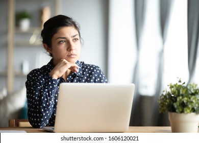 Serious thoughtful pensive young indian ethnic woman student sit at home office desk with laptop thinking of inspiration solution lost in thoughts dreaming looking away search creative ideas concept