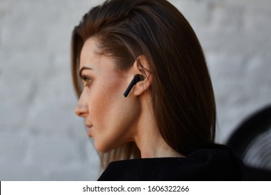 Young brown hair street fashion woman in black hoodie with opened neck turned back and uses to talk or listen music wireless earphones in ear in loft interior with white brick wall