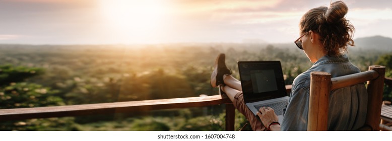 Mujer de negocios joven que trabaja en la computadora en el café en la roca. Desplazador de niña trabajando en una computadora portátil al atardecer o al amanecer en la cima de la montaña hasta el mar, día de trabajo.