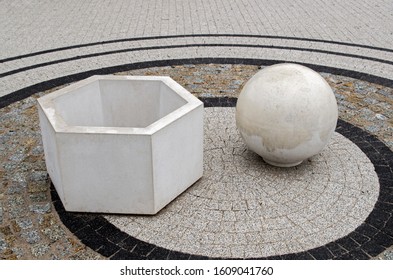 White concrete three-dimensional geometric shapes a hollow hexagon and a solid ball standing on a platform lined with gray, beige and black tiles within a black pavement circle