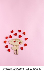Valentine's day. A wooden figure in the form of a couple under an umbrella is located on a paper pink background. Around the pair are small, wooden, red hearts. Flat lay composition with copy space.