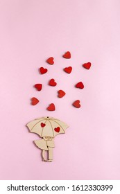 Valentine's day. A wooden figure in the form of a couple under an umbrella is located on a paper pink background. Around the pair are small, wooden, red hearts. Flat lay composition with copy space.