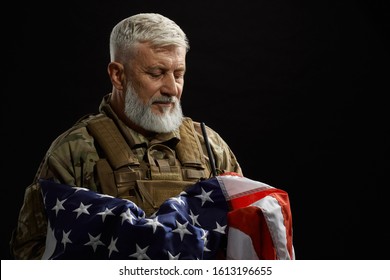 Front view of bearded american military veteran holding flag in arms. Portrait of old proud male officer in camouflage uniform posing in dark atmosphere. Concept of military, patriotism.