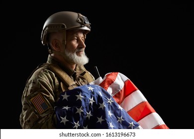 Front view of bearded american military veteran holding flag and looking away. Portrait of old male officer in camouflage uniform and helmet posing in dark atmosphere. Concept of military, patriotism.