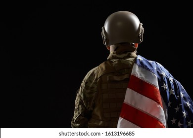Back view of american military veteran holding flag on shoulder. Portrait of old incognito proud male officer in camouflage uniform posing in dark atmosphere. Concept of military, army.