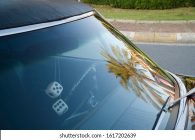 Detail jendela depan mobil Amerika klasik ungu di jalan. Ada ornamen mobil bergaya retro berbentuk dadu di kaca spion dan pantulan pohon palem di kaca depan.
