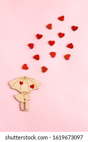 Valentine's day. On a paper pink background is located a wooden figure in the form of a pair under an umbrella. Around the pair are small, wooden, red hearts. Flat lay composition with copy space.