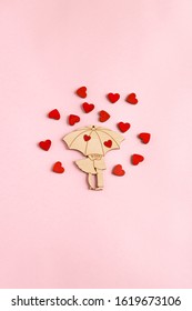 Valentine's day. On a paper pink background is located a wooden figure in the form of a pair under an umbrella. Around the pair are small, wooden, red hearts. Flat lay composition with copy space.