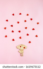 Valentine's day. On a paper pink background is located a wooden figure in the form of a couple under an umbrella. Around the pair are small, wooden, red hearts. Flat lay composition with copy space.