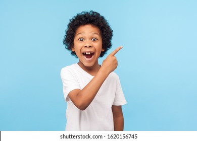 Wow schau, hier werben! Porträt des verblüfften niedlichen kleinen Jungen mit dem lockigen Haar, das auf leeren Platz auf Hintergrund zeigt, überraschter Vorschulkind, der Kopienraum für Werbeanzeige zeigt. Indoor-Studioaufnahme