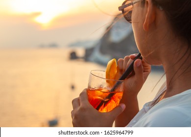 Mujer joven feliz con cóctel de naranja en bebida de vidrio al atardecer frente al mar en la costa de la isla de Ponza, en una pared con vista al océano. Sonriendo a la cámara. 9895