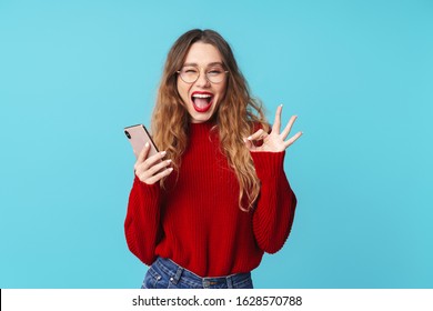 Imagen de una alegre mujer caucásica sosteniendo un teléfono celular y haciendo un gesto de ok mientras guiña un ojo aislado sobre fondo azul.