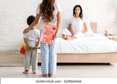 back view of brother and sister holding flowers and i love you mom card, and surprised woman sitting in bed
