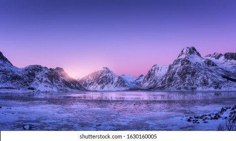 雪に覆われた山々、凍るような海岸のある青い海、水の反射、ノルウェーのロフォーテン諸島のカラフルな夕日の紫色の空。雪に覆われた岩のある冬の風景、夜は氷のフィヨルド。自然