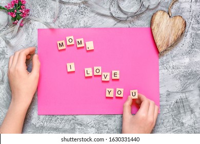 Happy Mothers Day Inschrift aus Holzbuchstaben auf rosa Hintergrund. Die Hände eines Kindes erfanden Worte für Glückwünsche an die Mutter. Symbole für Grußkartenvorlagen zum Muttertag.