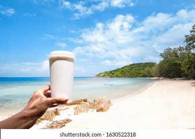 Mano sostiene una taza de papel de cartón blanco para bebidas para llevar, contra el hermoso fondo de la costa del mar, concepto de medio ambiente