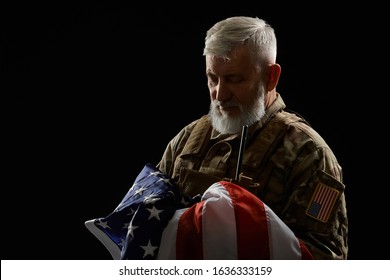 Front view of bearded american military veteran holding flag in arms. Portrait of old proud male officer in camouflage uniform posing in dark atmosphere. Concept of military, patriotism.