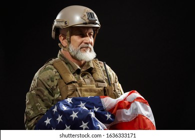 Front view of bearded american military veteran holding flag and looking away. Portrait of old male officer in camouflage uniform and helmet posing in dark atmosphere. Concept of military, patriotism.