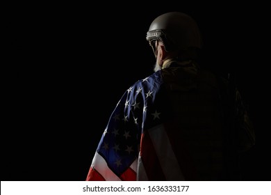 Back view of bearded american military veteran holding flag on shoulder. Portrait of old incognito proud male officer in camouflage uniform posing in dark atmosphere. Concept of military, army.
