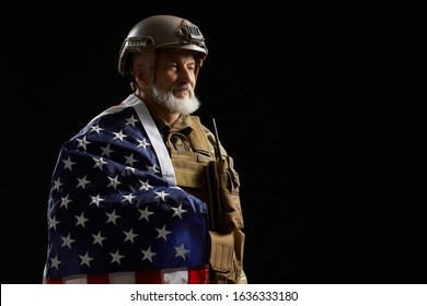 Side view of smiling american military bearded veteran holding flag on shoulder. Portrait of old proud male officer in camouflage uniform posing in dark atmosphere. Concept of military, army.