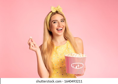Dag van de bioscoop. Portret van een opgewonden blond meisje dat popcorn eet en naar de camera kijkt, naar een film kijkt die over een roze muur is geïsoleerd