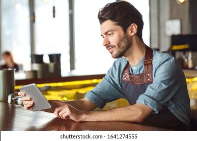 Professionele, bebaarde barman die op de toog staat en een digitale tablet vasthoudt die een online game speelt en vrolijk glimlacht
