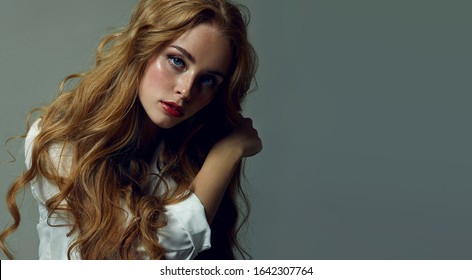 白い服を着てスタジオでポーズをとる豪華な長いウェーブのかかった赤い髪を持つ美しい青い目の少女。