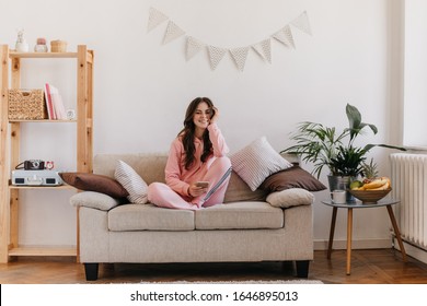 Beautiful brunette girl sitting on bright soft sofa surrounded by bookshelf and table with flower pot. Charming lady dressed in pink pajamas holds phone