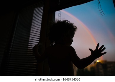 Kind met krullend haar bij het raam kijkt 's avonds naar de regenboog