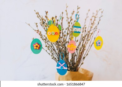 Telur Paskah dipotong dari kertas warna-warni yang dihias dengan pita dan kancing pada cabang pohon willow. Ide untuk dekorasi DIY bersama anak-anak. Fokus selektif pada telur kertas kuning.