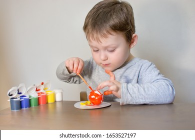 Cute, little beautiful child draws Easter eggs with colored paint. The boy is sitting at the table and enthusiastically engaged in creativity, preparing for Easter.