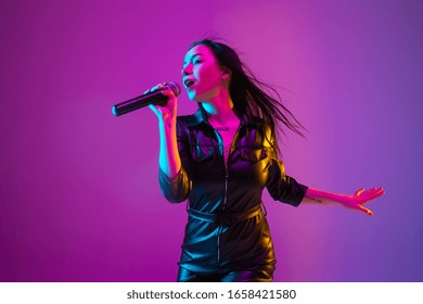 Caucasian female singer portrait isolated on purple studio background in neon light. Beautiful female model in black wear with microphone. Concept of human emotions, facial expression, ad, music, art.