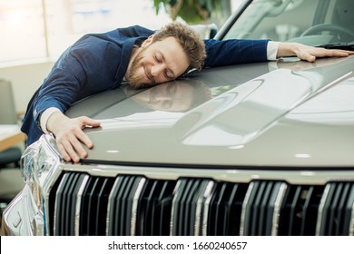 ディーラーで新車を抱きしめる正式なスーツを着た面白い若い白人のハンサムな男。新しい自動車を買った後の幸せな陽気な男