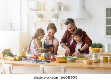 キッチンで一緒に料理をする幸せな家族。父と息子にレシピを読んでいる母と娘。お父さんと男の子がサラダ用に緑の野菜の葉を刻んでいます。週末の家庭でのレクリエーションと食事の準備